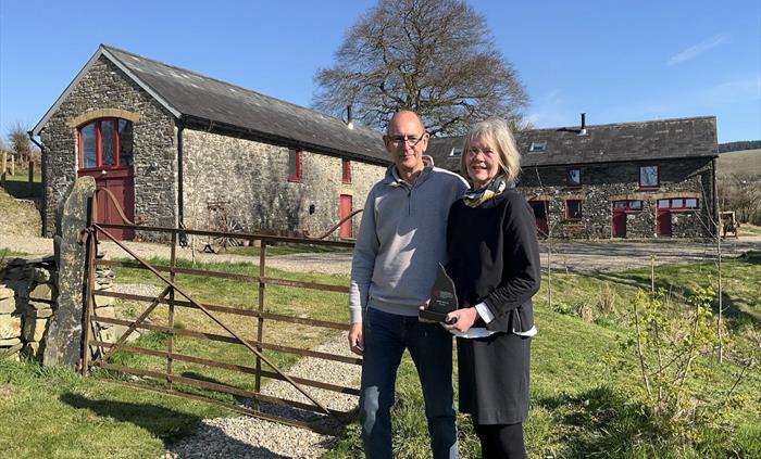 A view of the owners in front of the barn, holding their 2025 National Tourism Award for Wales 'Bro a Byd' trophy